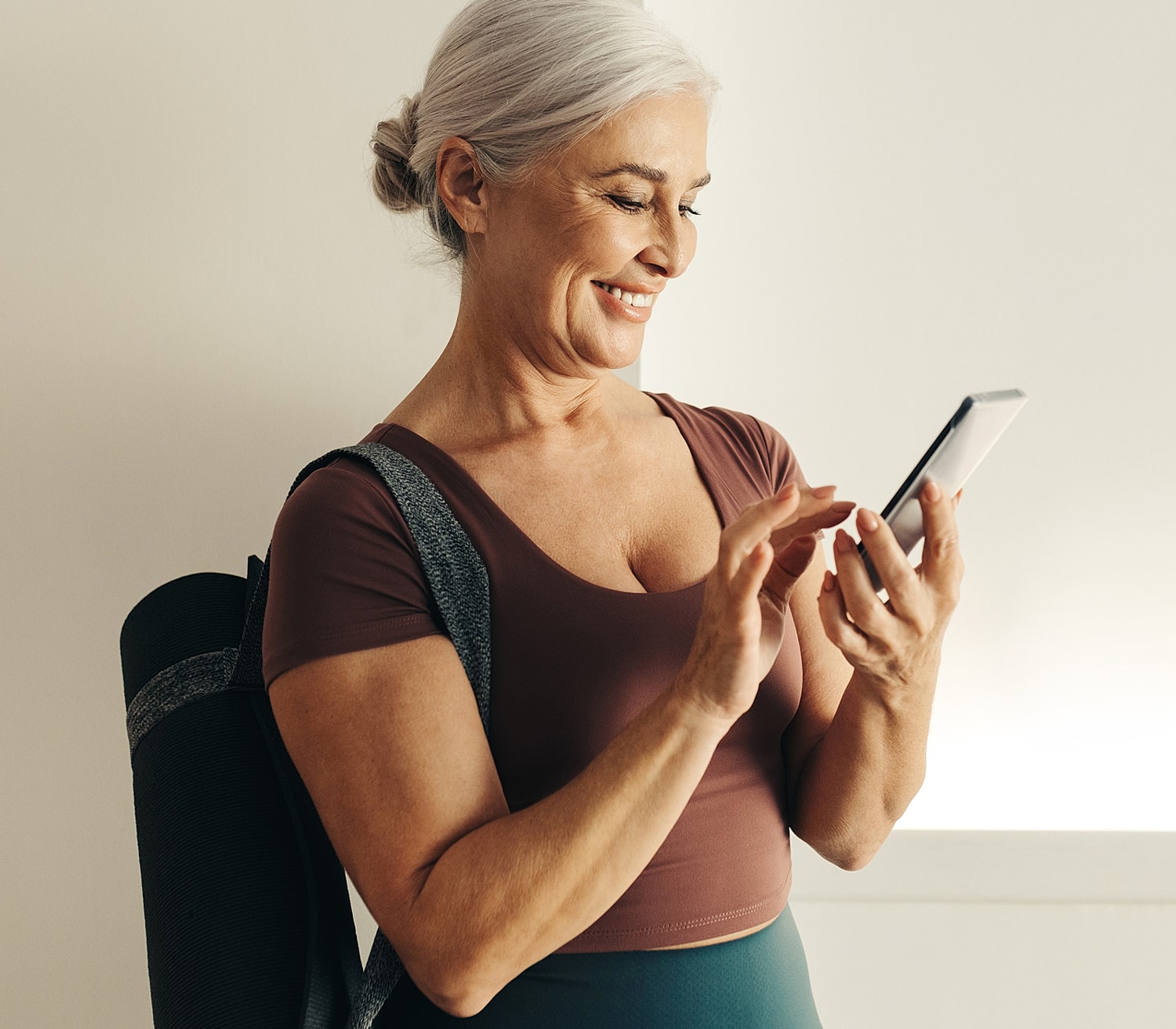 Smiling woman checking phone with yoga mat.